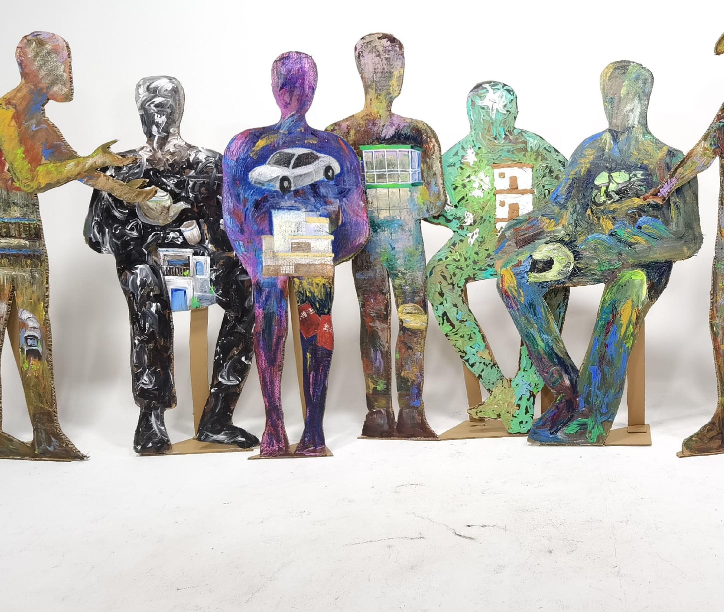 Painted cardboard figures carrying a white car, an apartment window, a house, and a stack of banknotes across their bodies