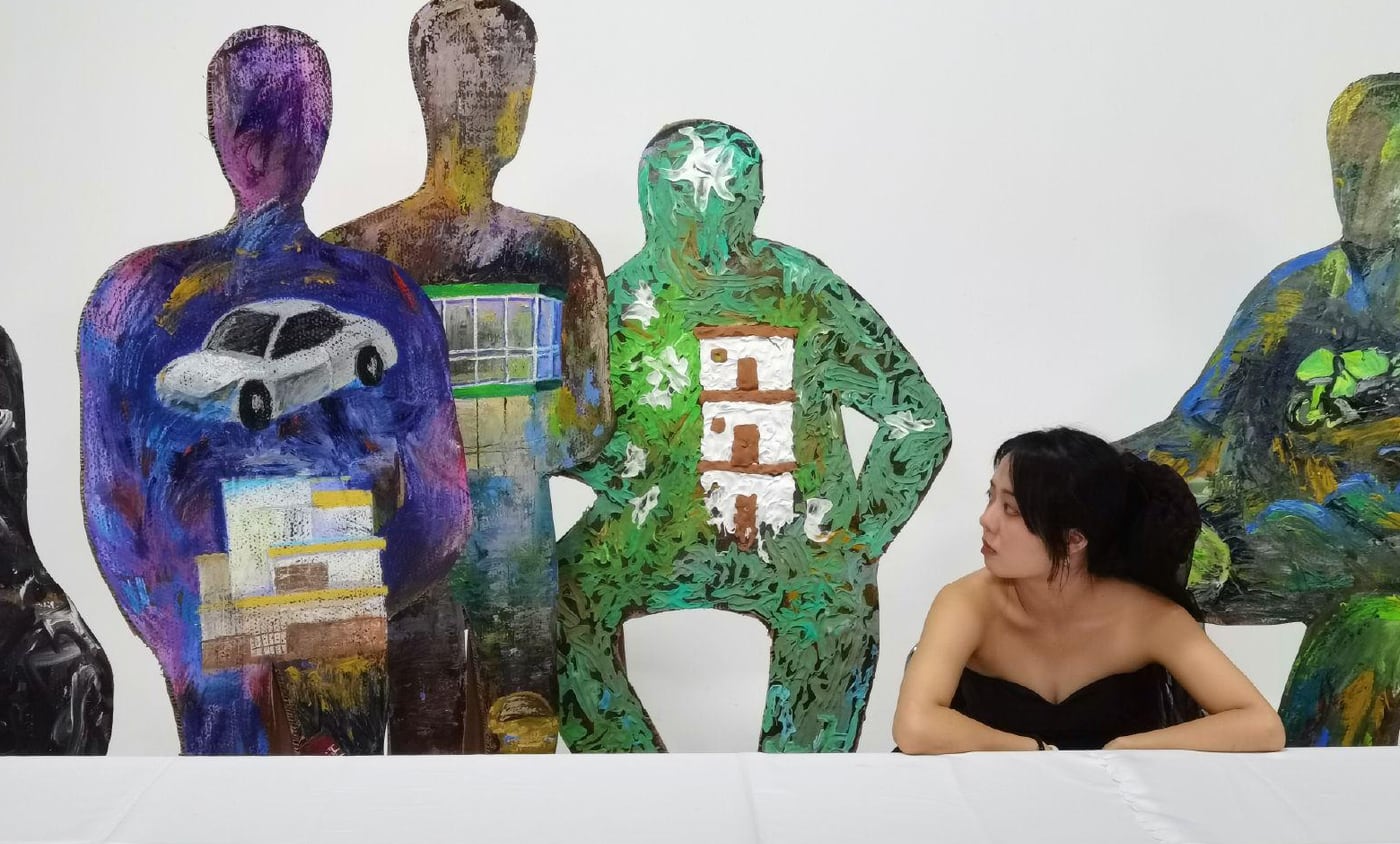 The artist seated at the long white table, turning to look at the painted cardboard figures beside her