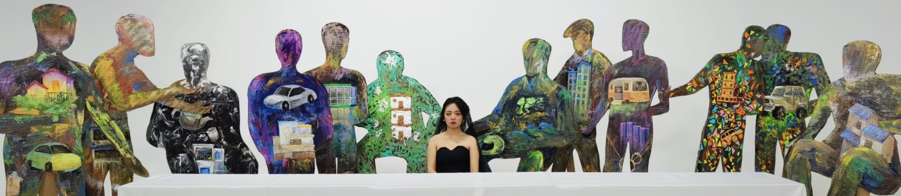 The twelve painted cardboard figures ranged left and right behind a long white banquet table, with the artist seated at the centre