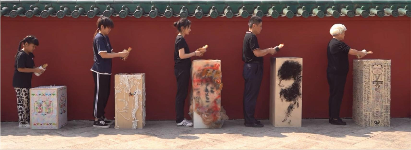 Five performers of different ages stand along a red temple wall, each beside their own mixed-media pillar