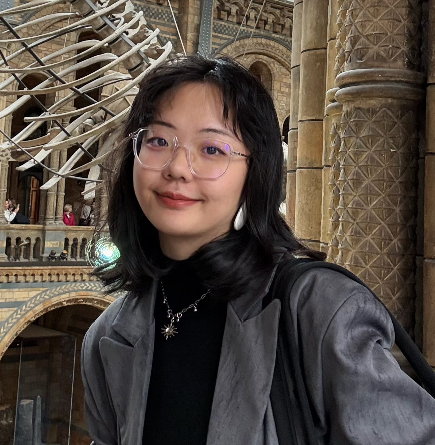 Portrait of Mengfan He, wearing glasses and a grey jacket, in a museum hall with a whale skeleton and carved stone columns behind her